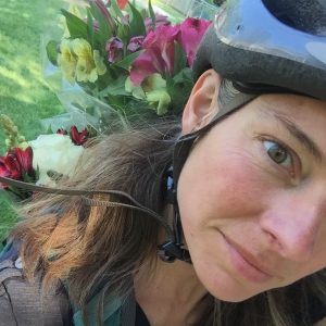 Susan is dressed in concert black and wearing a bike helmet. She is carrying a backpack with flowers, which were a thank you gift from a performance, sticking out of the back. Photo by the author ca. 2017.