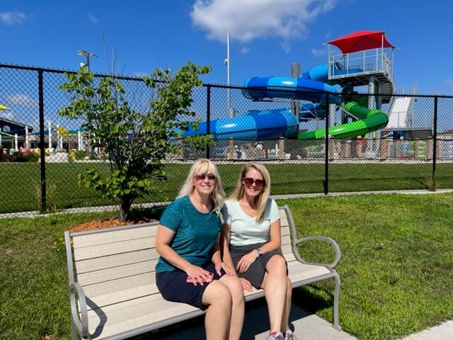 Dawn Desorcy (Parks & Recreation Coordinator) and Carrie Edmonson (Associate Planner) sit on a bench at Witter Park. Behind them is the new aquatic center.